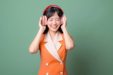 Güneydoğu Asyalı, turuncu elbiseli, gözleri kapalı gülümseyen yeşil arka planda kulaklıkla müzik dinleyen bir kadın.