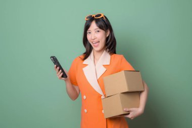 Turuncu elbiseli mutlu bir kadın, yeşil arka plan, dağıtım ve ticaret kavramına karşı karton kutular ve telefon taşıyor.