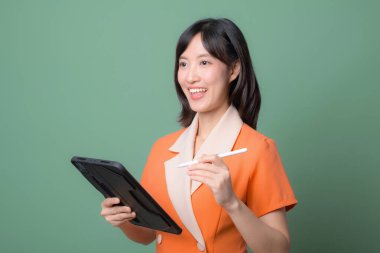 Güneydoğu Asyalı bir kadın elinde dijital tablet ve stilini yeşil arka planda tutarken gülümsüyor, modern profesyonel ortamda mutluluk ve güveni ifade ediyor.