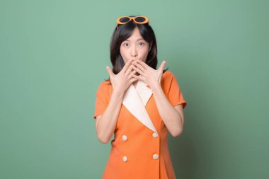 Kafasında güneş gözlüğü olan, sürpriz bir güneydoğu Asyalı kadın yeşil arka plana karşı duruyor. Ağzını kapayarak, ellerini şokla gösteriyor.