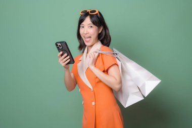 Turuncu elbiseli güneydoğu Asyalı bir kadın alışveriş torbaları tutuyor ve mutlu bir ifadeyle akıllı telefon kullanıyor. Yeşil arka planda internet alışverişi kavramı.