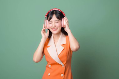 Güneydoğu Asyalı, turuncu elbiseli, kulaklıkları kapalı, yeşil arka plana gülümseyen bir kadın müzik dinliyor.