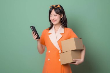 Turuncu elbiseli, elinde karton kutular olan güneydoğulu bir kadın yeşil arka planda durup gülümseyen akıllı telefon kullanıyor.