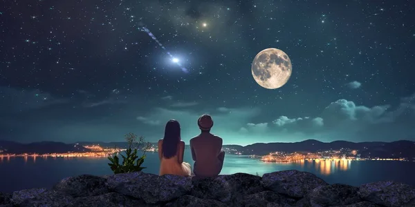 young couple man and woman sit on rock at night sea watching starry sky and big moon on horizon mediterranean city blurred light reflection on water romantic wallpaper
