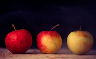 Elmaların önü koyu arkaplan görünümlü ahşap bir masada üç elma.