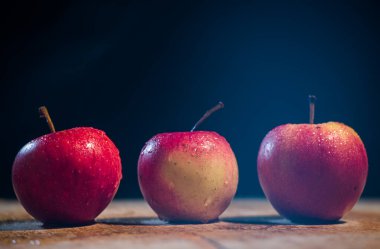 Elmaların önü koyu arkaplan görünümlü ahşap bir masada üç elma.