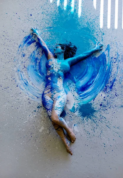 白で若いセクシーな裸の女性 青とターコイズ色の創造的な 抽象的な塗装 嘘の装飾 アーティストのスタジオの床の上にエレガントな装飾 ブリーチ地面に太陽の光の平行線 スペースをコピー — ストック写真