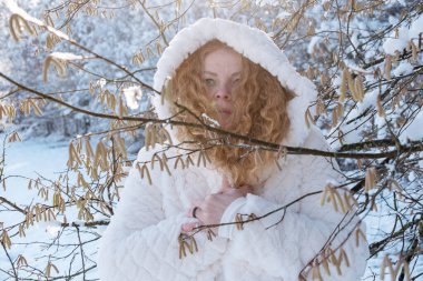 portrait of beautiful sexy mature redhead woman, with red curly hair in white hooded coat in winter nature sunlight, snowy tree, copy space