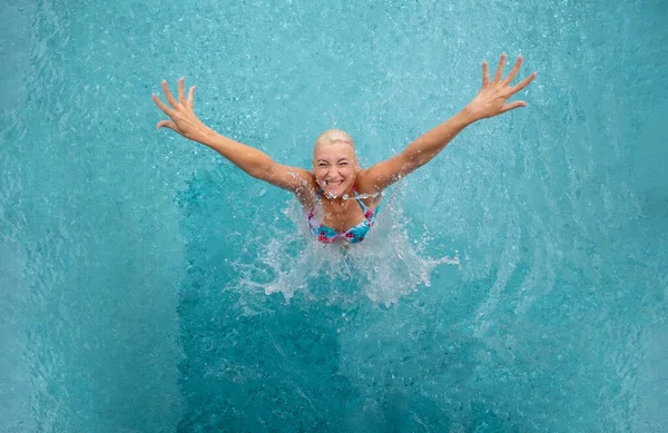 top view to young sexy blonde happy laughing bikini woman in turquoise pool jumps up from splashing water with outstretched arms, copy space