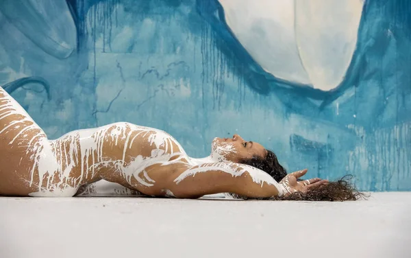 portrait and upper body, torso of sexy nude brunette woman, long hair, curly, artistically abstract painted, body covered with white paint, lies on the studio floor under lilly painting, copy space
