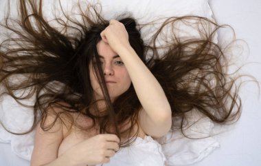 Çekici, genç, seksi, esmer bir kadın, saçı çarşafların üzerinde vahşi, doğrudan bakan, saçlarıyla oynayan, gevşemiş, yatakta beyaz çarşafların içinde uzanan,