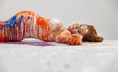 Genç, seksi, baştan çıkarıcı, büyük yuvarlak göbekli, turuncu, mavi, beyaz, sanatsal soyut resimli, sanatçı stüdyosunda, renkli zemine uzanmış,