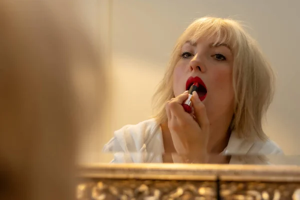 Blonde woman stands at the bathroom mirror and puts on makeup and lipstick, copy space