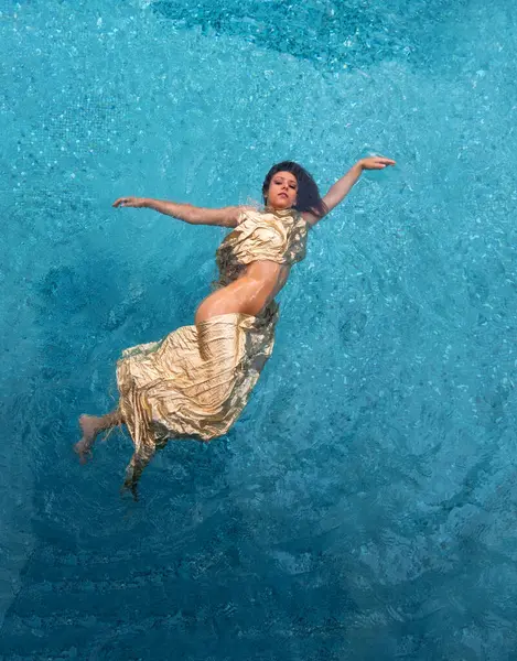 top view to beautiful young sexy nude brunette woman in fashion golden dress, swimming floating weightless elegant in the turquoise water in the pool, copy space