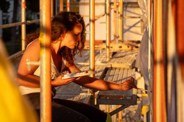 Kıvırcık esmer saçlı seksi kadın ressam sokak sanatını boyuyor iskele üzerindeki duvar resmini fırça ve palet ile silikleştiriyor sıcak gün batımı ışığında ön cephede, fotokopi uzayı
