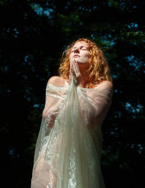 Şehvetli, baştan çıkarıcı, kızıl saçlı Madonna portresi, dua ederken katlanmış eller, kapalı gözler, parlak şeffaf kumaşa sarılmış çıplak..