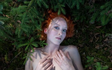 Seksi kızıl saçlı bir kızın portresi orman zemininde küçük çam ağaçları arasında yüzüyor.