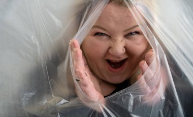 mischievously maliciously laughing woman sticks her head and hands out of the plastic foil wrap, like a witch from a fairy tale, copy space clipart