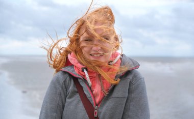 Kızıl saçlı bir kadın rüzgarlı bir sahilde duruyor, saçları çılgınca uçuşuyor. Gri bir ceket ve pembe bir kapüşonlu ceket giyiyor. Kasvetli havaya rağmen gülümsüyor. Arka plan bulutlu bulutlu bir gökyüzünü gösteriyor.