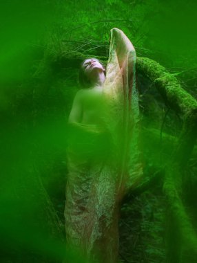 Kadın yemyeşil bir ormanda duruyor, kısmen şeffaf kumaşa sarılmış, yosun kaplı bir dala yaslanmış, yukarı doğru bakıyor, sahne ruhani ve canlı, kopyalama alanı