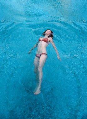 Mavi bir havuzda, bikini giymiş bir kadın huzur içinde yüzüyor. Güneş ışığı suyun üzerinde dans eder, dingin ve ferahlatıcı bir atmosfer yaratır. Rahatlık ve sükuneti somutlaştırıyor..
