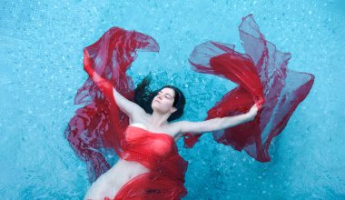 Bir kadın canlı kırmızı kumaşla örtülü bir havuzda huzur içinde yüzüyor. Kırmızının mavi sudaki zıt renkleri çarpıcı, huzurlu ve zarif bir manzara yaratır..