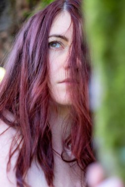 Uzun kızıl saçlı bir kadın kısmen yüzünü kapatıyor, mavi gözleri ipliklerden içeri bakıyor. Yakın plan, yoğun, derin düşüncelere dalmış ifadesini doğal arka planla harmanlıyor..