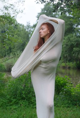 Seksi kadın, vücudunu çevreleyen şeffaf beyaz bir kumaşa sarılmış olarak doğada duruyor. Kumaş kafasının üstüne doğru uzanıyor, gerçeküstü ve ruhani bir etki yaratıyor, ifade derin derin düşünüyor.