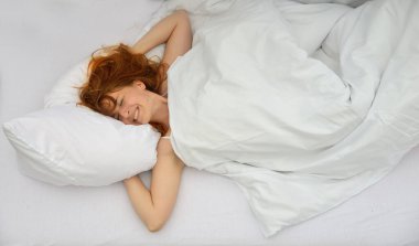 Yatakta yatan seksi kızıl saçlı kadın, gözleri kapalı gülümsüyor, beyaz bir battaniyeyle örtülmüş ve beyaz bir yastığın üzerinde dinleniyor, huzurlu ve rahat bir atmosfer yaratıyor, boşluğu kopyalıyor.