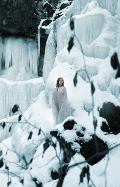 Saf beyaz kumaşa sarılı seksi genç bir kadın donmuş bir kış manzarasının ortasında duruyor. Buz saçakları ve karla kaplı kayalar dingin ve ruhani bir manzara yaratıyor.
