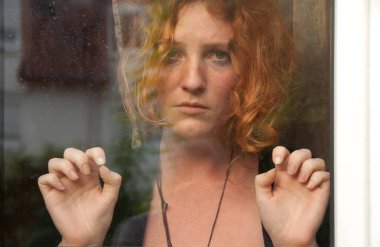 Seksi, genç, kızıl saçlı bir kadın ellerini cama dayayıp derin düşüncelere dalmış bir anda özlem, üzüntü ve duygusal derinlik hissi uyandırıyor.