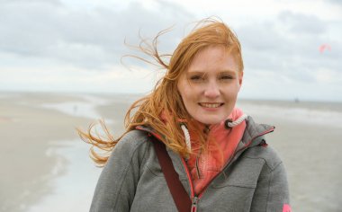 Kızıl saçlı kadın esintili bir plaj arka planına sıcak bir şekilde gülümsüyor, saçları rüzgarda dans ediyor. Bulutlu gökyüzü ve sakin kıyı şeridi bir sakinlik hissi uyandırıyor ve doğayla bağlantı kuruyor, boşluğu kopyalıyor