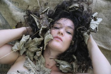 Sexy brunette woman lies surrounded by dried autumn leaves on a burlap surface, her dark curly hair blending with the natural textures. Her serene expression evokes a connection to nature, copy space clipart