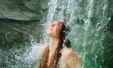 young sexy redhead woman stands under a cascading waterfall, her face tilted upwards in serene joy. The rushing water, her peaceful expression evoke freedom and pure connection to nature, copy space clipart