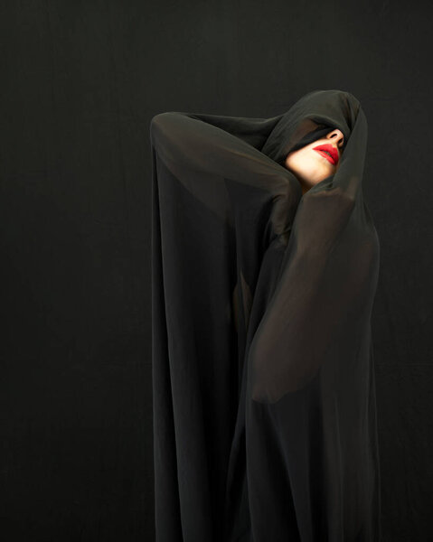 сексуальная молодая загадочная женщина, покрытая черной тканью, ее красные губы смело контрастируют с темнотой, вызывает загадочную и элегантную ауру в этом минималистичном и драматичном портрете, копирующем пространство