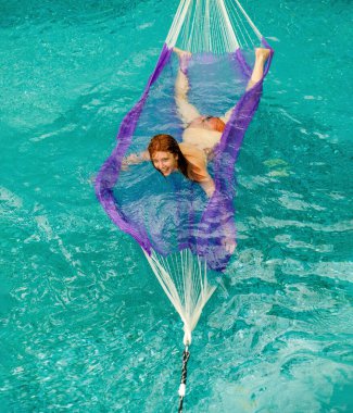 Neşeli kızıl saçlı kadın ve bir erkek neşeli bir şekilde canlı bir turkuaz havuzda yüzen eşsiz bir hamağın keyfini çıkarıyorlar. Gülüşleri ve hareketleri eğlenceli, kaygısız ve ferahlatıcı bir yaz havası yaratıyor.
