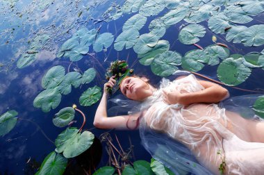 Seksi baştan çıkarıcı kadın su zambaklarıyla çevrili bir gölde yüzüyor ve Ophelia 'nın trajik güzelliğini hatırlatıyor. Saf kumaşa sarılmış, elinde çiçeklerden bir taç var, rüya gibi bir durumda kaybolmuş, boşluğu kopyalıyor.