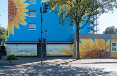 Canlı bir ayçiçeği duvar resmi şehir manzarasına sıcaklık ve neşe getirir. Parlak sarı çiçekler mavi duvarla güzel bir tezat oluşturuyor, neşeli ve sanatsal bir atmosfer yaratıyor..