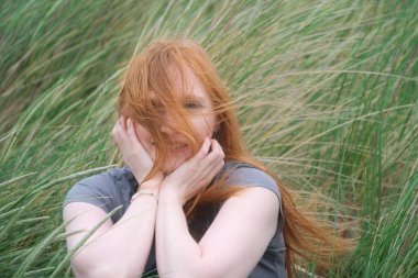 Uzun sahil çimlerinde canlı kızıl saçlı bir kadın. Rüzgâr saçlarını dalgalandırıyor. Bir neşe, özgürlük ve doğayla bağlantı anı, sıcaklık, hareket ve doğallık ile dolu, kopyalama alanı