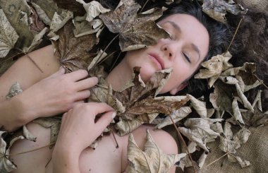 Duygusal, şehvetli, baştan çıkarıcı, seksi, kadın zarif bir şekilde sonbahar yaprakları arasında uzanıyor, koyu esmer, kıvırcık saçlar doğal dokularla harmanlanmış, samimi sanatsal bir hava yaratıyor, boşluğu kopyalıyor.