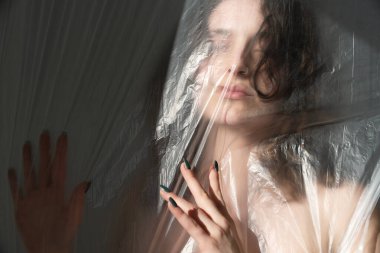 Şeffaf plastiğin arkasındaki şehvetli kadın, elini uzatıyor, yüzü bulanık ve bozuk. İzolasyon, kırılganlık, hasret, incinebilirlik ve gizli duygular için görsel bir metafor.