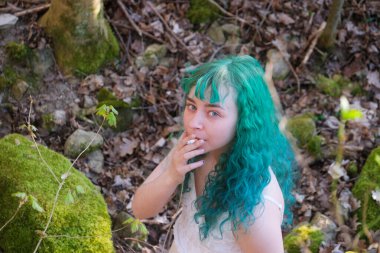 Canlı turkuaz yeşil saçlı, beyaz elbiseli, yosunlu orman ortamında sigara içen genç bir kadın. Doğal, asi ve rüya gibi bir atmosfer..