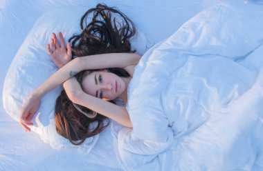 Yumuşak bir sabah ışığında beyaz bir battaniyenin altında uzanmış, gevşemiş, sakin bir ifadeyle esneyen seksi genç kadın. Huzurlu, rüya gibi bir ruh hali. Yastığın üzerine koyu renk saçlar yayılmış. Boşluğu kopyala.