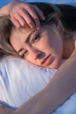 Seksi genç kadın beyaz bir yastığa uzanır, yüzü ılık sabah güneşiyle hafifçe aydınlanır. Onun gevşek ifadesi, dağınık saçları ve nazik dokunuşu huzurlu, samimi bir yakınlaşma anı yaratır..