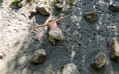 Güçlü bir kadın Viyana 'da Tuna Nehri yakınlarında diz çöker, kollarını kumlu zemine uzatır. Kayalar ve ışıkla çevrili duruşu ritüel gücünü ve doğayla ham bağlantısını anımsatıyor.