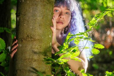 Leylak saçlı genç bir kadın, parlayan bir yaz ormanında bir ağacı kucaklıyor, elleri yavaşça kabukta dinleniyor, taze yeşil yapraklar ve yumuşak güneş ışığı ile çevrili..