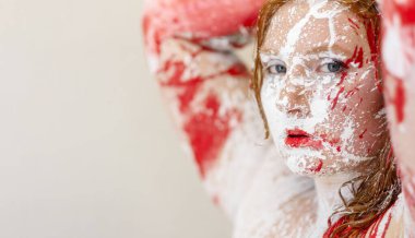 Yoğun mavi gözlü, yüzü ve vücudu kalın kırmızı ve beyaz boyayla kaplanmış cesur, ham ve resmî bir duygu ifadesi yaratan genç bir kadının yakın çekimi..