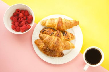 Kahve fincanı, taze kruvasan ve ahududu sarı ve pembe arka planda.