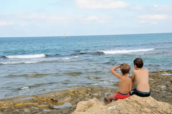 Zwei Jungen Sitzen Auf Küstenfelsen Und Betrachten Eine Schwimmende Grenzboa lizenzfreie Stockbilder