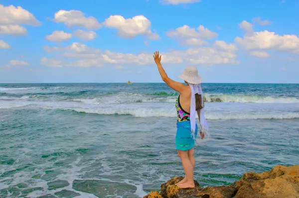 一个戴着白帽的女孩站在岩石上 向在海里航行的船只挥挥手 图库图片